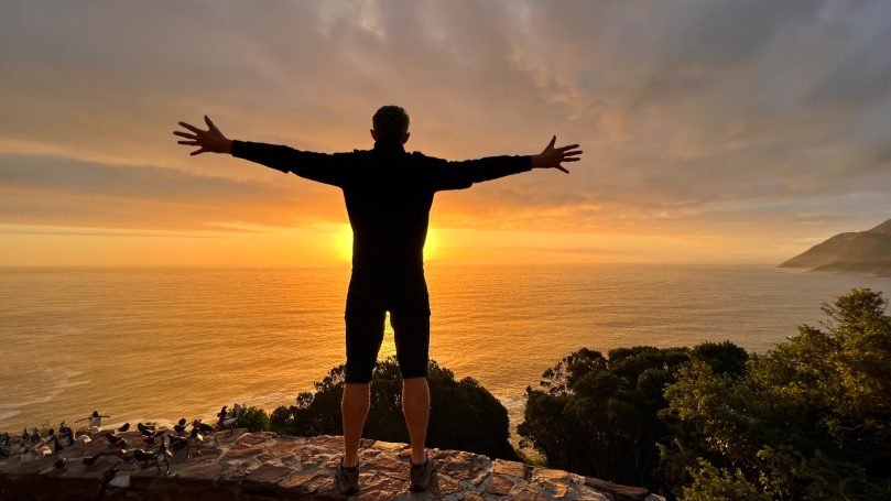 Mann mit erhobenen Armen vor Sonnenuntergang über dem Meer.