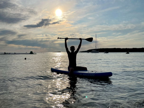 Person auf einem Stand-Up-Paddleboard hebt ein Paddel gegen die Sonne.