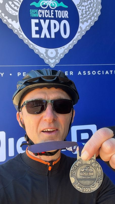 Radfahrer mit Sonnenbrille hält eine Medaille vor einem blauen Hintergrund.